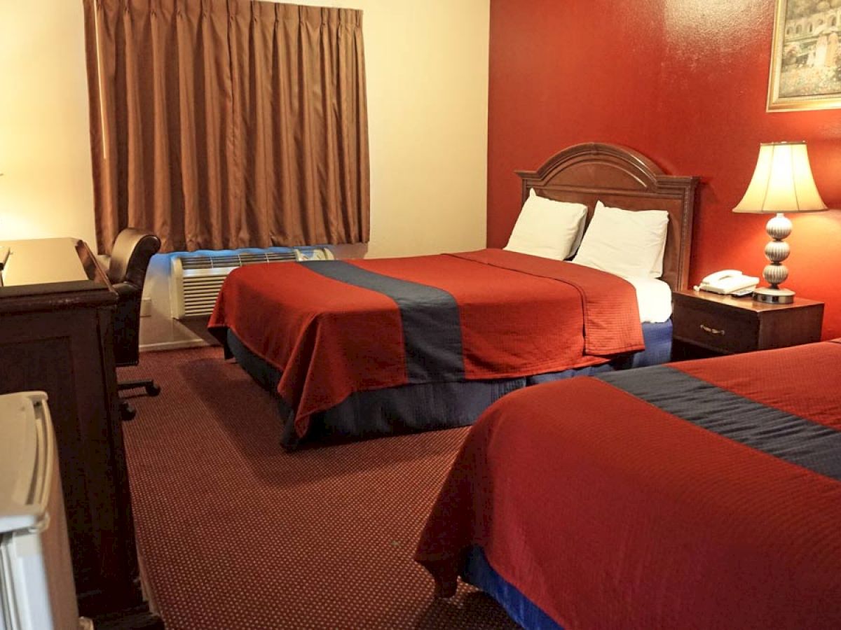 A hotel room with two double beds, red and blue bedding, a TV, desk, lamp, telephone, and brown curtains adorn the window on a red accent wall.