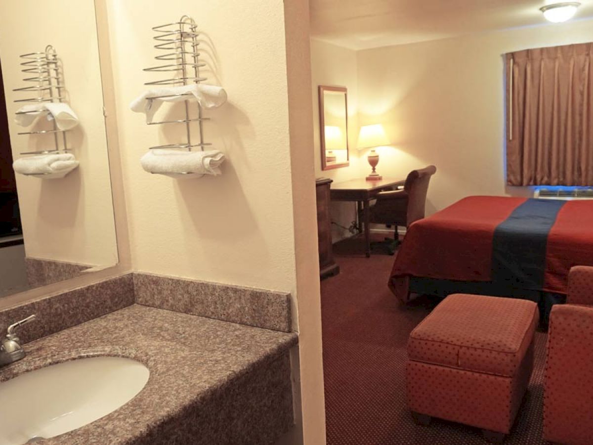 A hotel room with a sink and towels on the left, a bed, a desk with a chair, a lamp, an ottoman, and a curtain covering the window on the right.