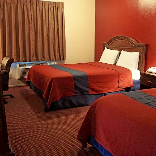 This image shows a hotel room with two double beds, a nightstand with a lamp, a TV on a desk, a chair, and a window with brown curtains.