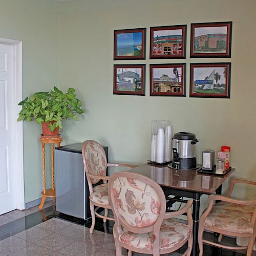 A small room with a table and four chairs, a coffee maker, a green plant, and framed pictures on the wall is shown in this image.