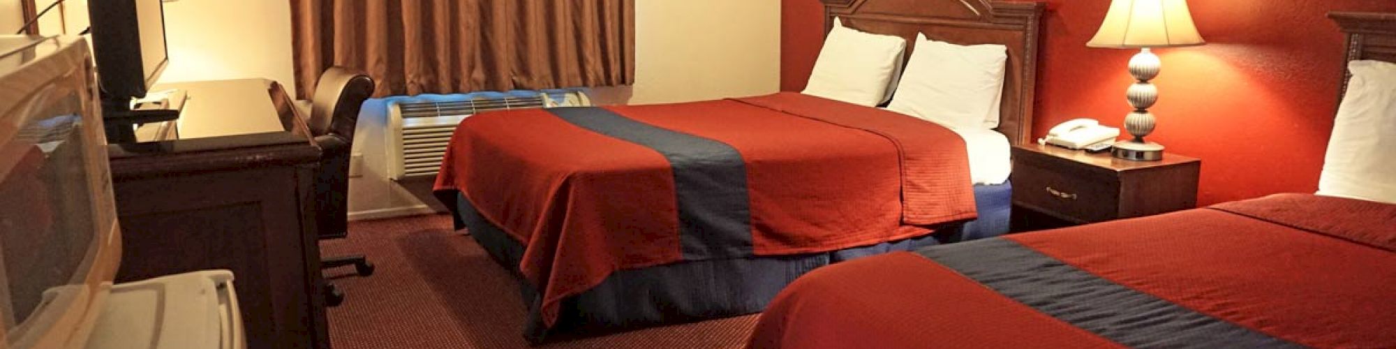 A hotel room with two double beds with red and blue bedspreads, a lamp, a TV, a desk, a microwave, and a window with brown curtains.