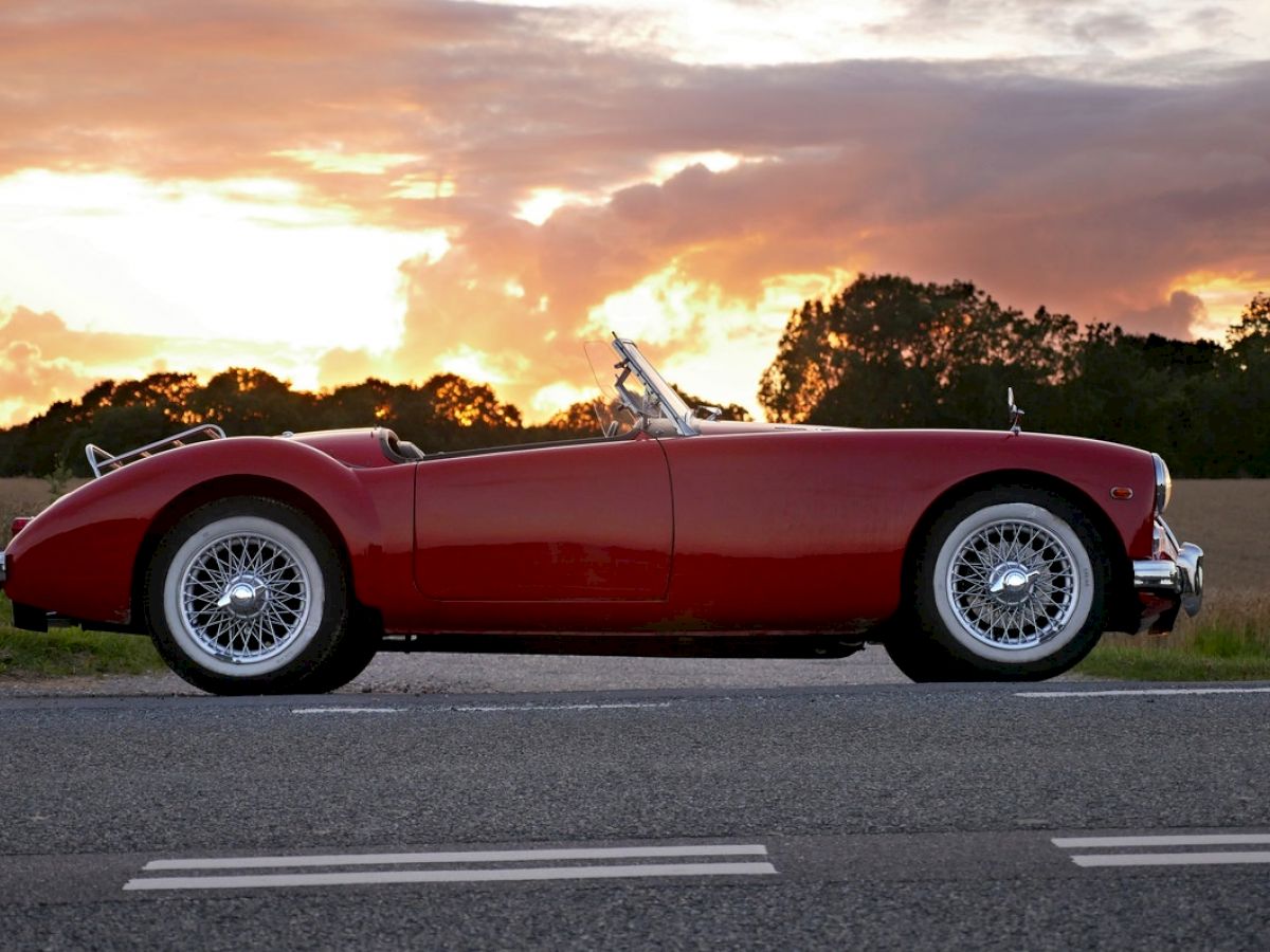 A classic red convertible car is parked on the side of a road during sunset, with a scenic background of fields and trees.