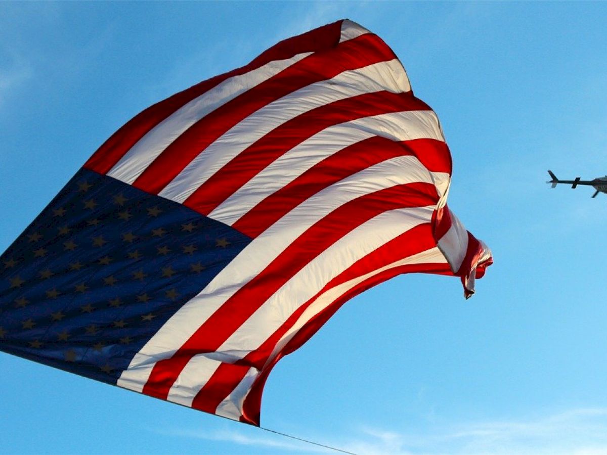 An American flag waves in the wind against a blue sky, with a helicopter flying nearby in the background.