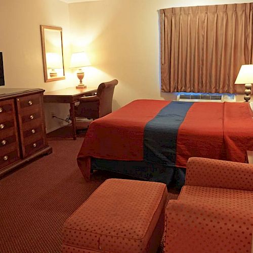 The image depicts a hotel room with a queen-sized bed, red and blue bedding, a TV on a dresser, a work desk, a chair, and multiple lamps.