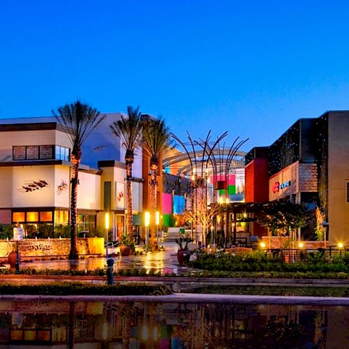 The image shows a brightly lit outdoor shopping and entertainment center at dusk, with palm trees and a variety of stores and restaurants.