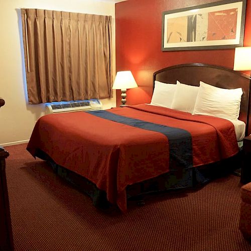 A hotel room with a king-sized bed, red and blue bedding, two lamps, a window with curtains, TV, armchair, and art on the walls.