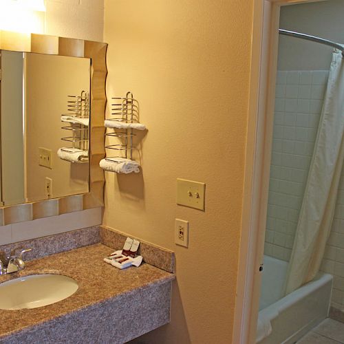 The image shows a bathroom with a mirror, sink, and towels on a shelf. To the right, there's a toilet and a bathtub with a shower curtain.