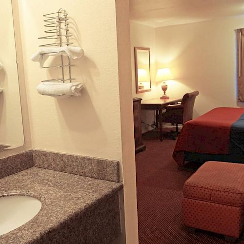 The image shows a hotel room with a vanity sink area, a bed, chairs, a desk with a mirror, lamps, and a window with curtains.