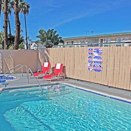 The image shows an outdoor swimming pool with two red lounge chairs, a wooden fence, and a sign on the fence. Palm trees and a building are visible in the background.