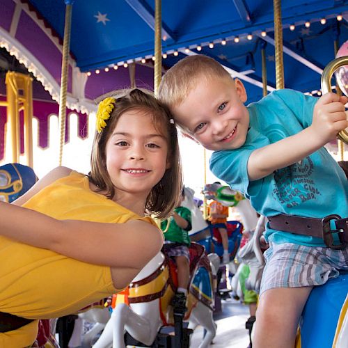 Two children are enjoying a ride on a brightly colored carousel, smiling happily while holding onto the poles.