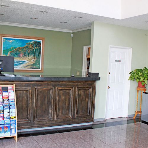 A reception area with a wooden desk, computer, brochures display, door, chairs, and framed pictures on the walls, plus a potted plant.