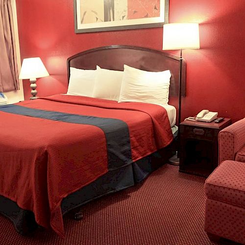 This image shows a cozy hotel room with a red theme, featuring a large bed, two lamps, an armchair, a nightstand, and a painting on the wall.