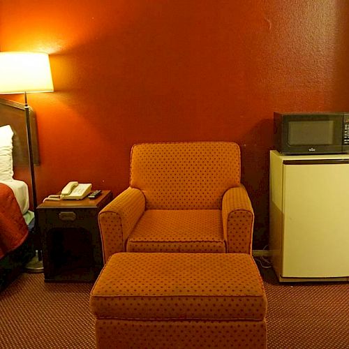 A hotel room with a bed, nightstand, lamp, chair with ottoman, mini fridge, microwave, and an open closet with an ironing board and iron.