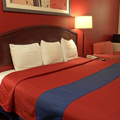 A well-lit hotel room with a large bed, red and blue bedding, lamps, a nightstand, a chair, and a small adjacent bathroom visible in the background.