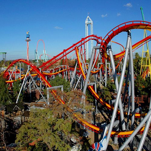 The image shows a theme park with several roller coasters, featuring bright orange and yellow tracks, surrounded by trees and other rides ending the sentence.