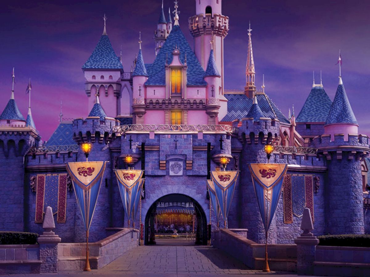 A grand castle with blue rooftops and flags sits illuminated against a twilight sky, inviting visitors through its arched gateway.