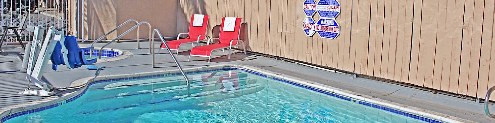 A swimming pool area with clear blue water, red lounge chairs, safety signs on a wooden fence, and palm trees in the background.