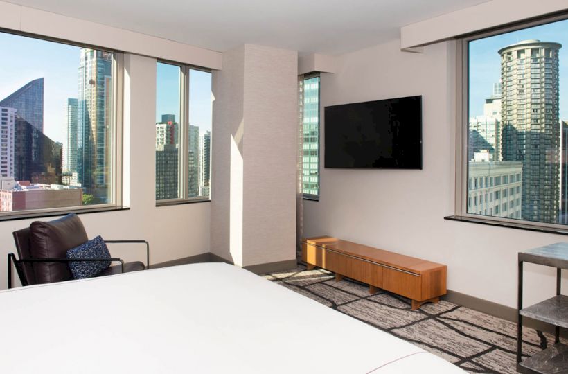 A modern hotel room with large windows, city view, bed, leather chair, wall-mounted TV, and a wooden console.