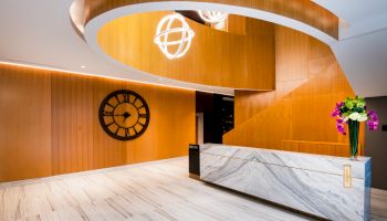 The image shows a modern lobby with a marble reception desk, a large decorative clock on the wall, and stylish hanging lights above.
