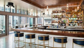 The image shows a modern bar interior with five barstools, a well-stocked bar, and large windows offering a view of the city.