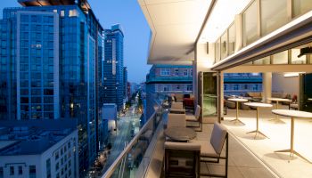 The image shows a modern rooftop balcony overlooking a cityscape at dusk, featuring outdoor seating and tables adjacent to indoor spaces.