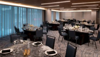 The image shows a modern banquet hall set up with round tables and chairs, neatly arranged with table settings, floral centerpieces, and dim lighting.