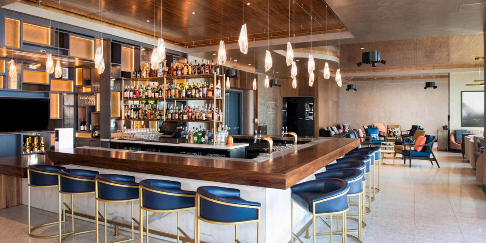 The image features a modern bar with blue barstools, a well-stocked liquor display, hanging light fixtures, and a seating area with sofas and chairs.