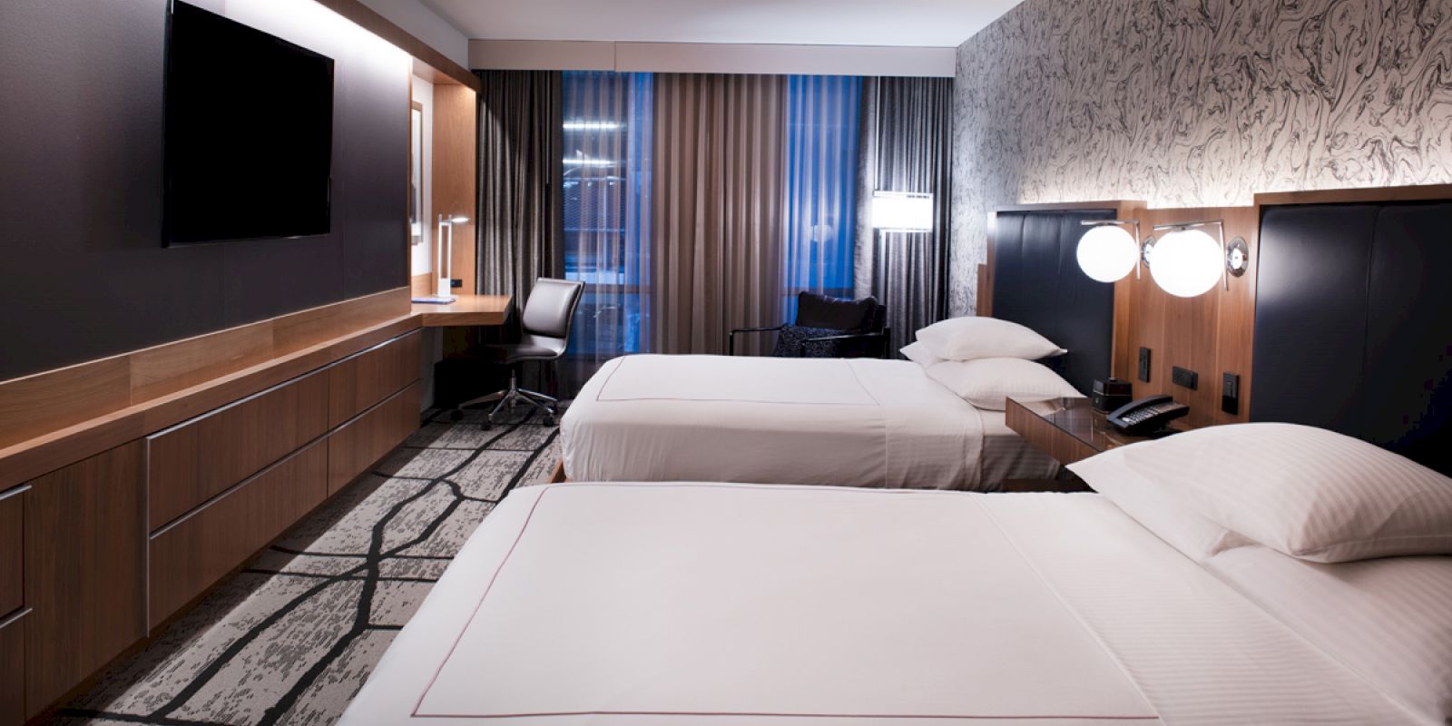 A modern hotel room features two beds, a flat-screen TV, a desk with a chair, a window with curtains, and ambient lighting.