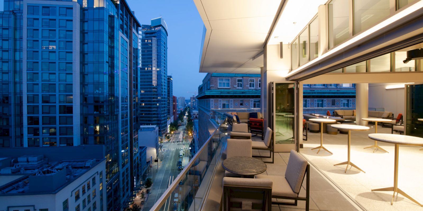 The image shows a modern cityscape at dusk with tall buildings and a lit, sleek balcony with tables and chairs overlooking a street.