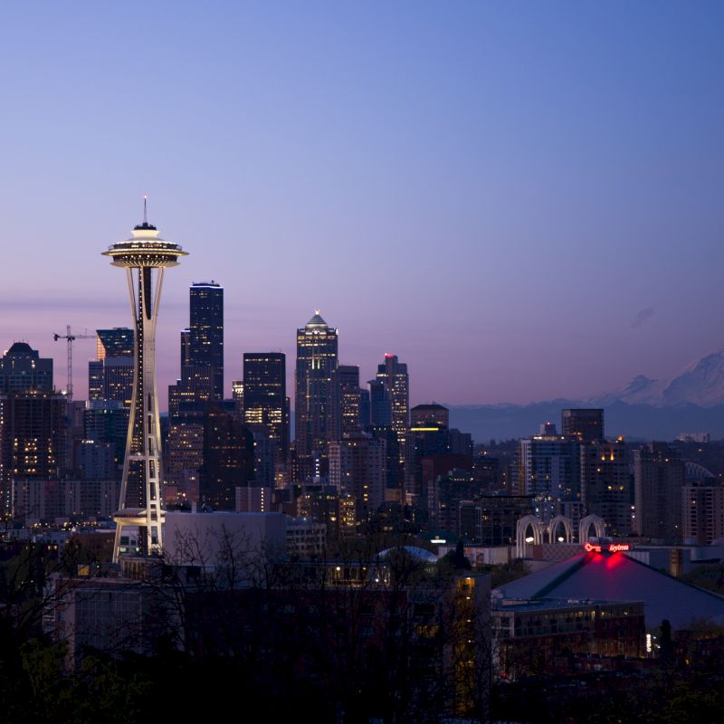 The image shows a cityscape with the Space Needle in the foreground, surrounded by skyscrapers, and a mountain visible in the background at dusk.
