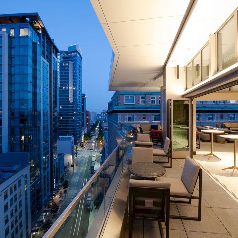 A modern, open-air balcony with tables and chairs overlooking a cityscape at dusk, with tall buildings and illuminated streets below.