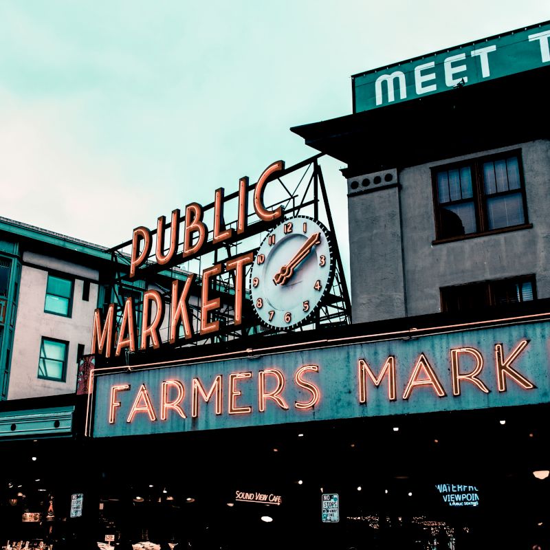 The image shows the entrance to a farmer's market with a clock under neon signs reading "Public Market" and "Farmers Market," and surrounding buildings.