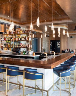 This image shows a modern bar with blue chairs, pendant lights, and a fully stocked bar. Seating and furniture are also visible in the background.