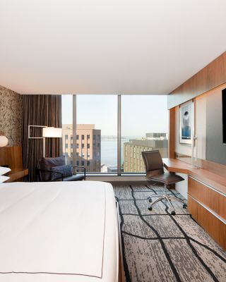 A modern hotel room features a large bed, a desk, a chair, and a flat-screen TV. The window offers a view of buildings and water outside.
