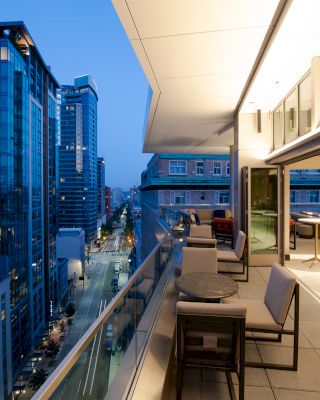 A modern city view with tall buildings at dusk, featuring a spacious balcony with tables and chairs lit by warm lighting.