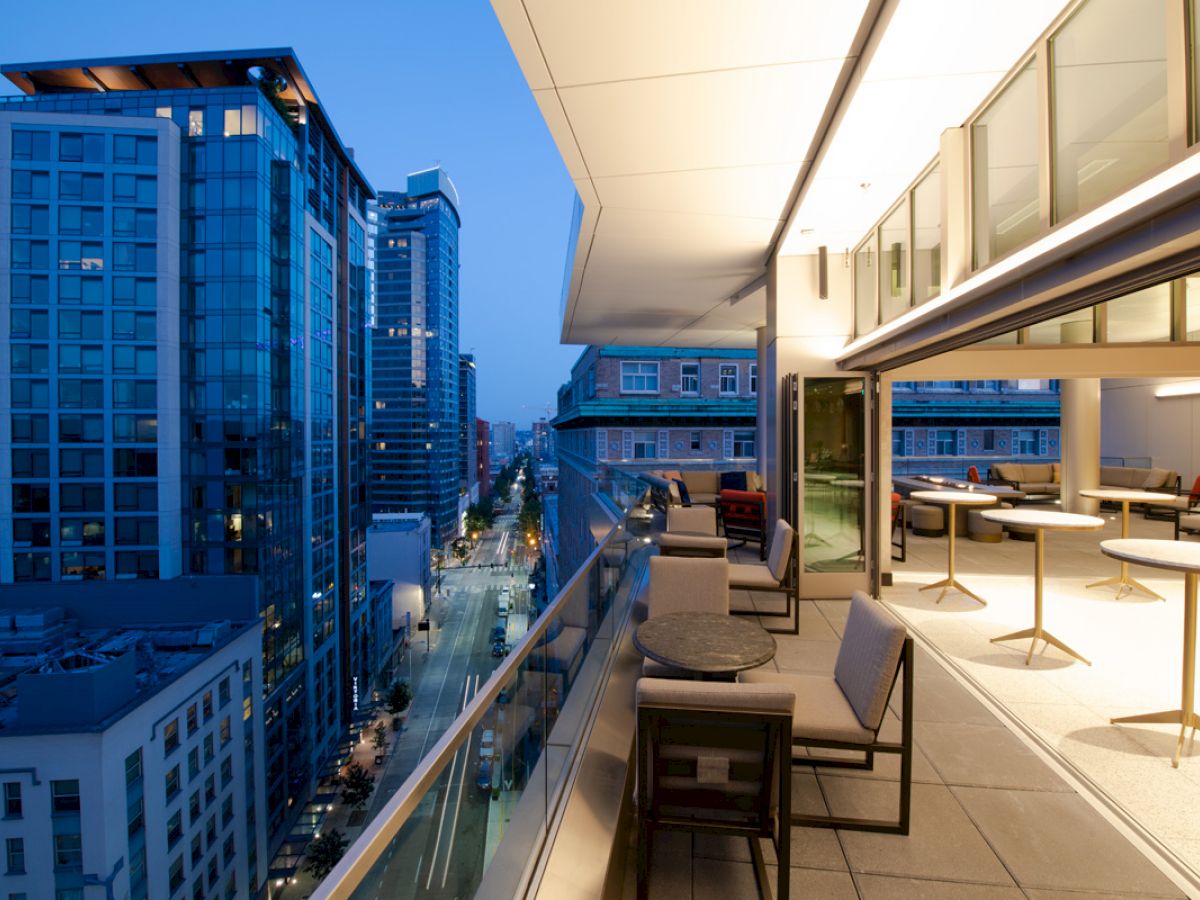 A modern outdoor balcony at dusk, showcasing a cityscape view with tall buildings, streetlights, and a sleek, illuminated seating area.