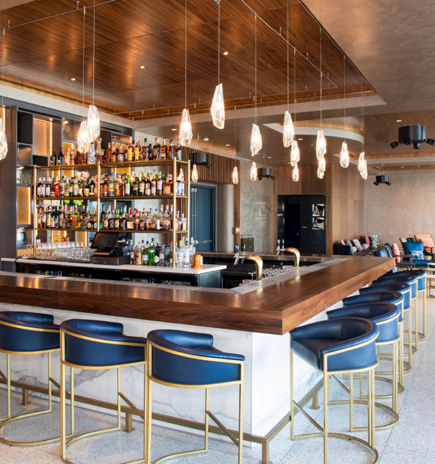 Modern bar with hanging lights, a central counter, bar stools, a variety of alcohol bottles, and seating area in the background.