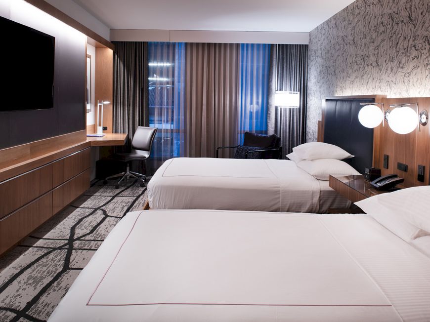 A modern hotel room with two beds, a desk with a chair, a flat-screen TV, and contemporary lighting and decor, featuring large windows.
