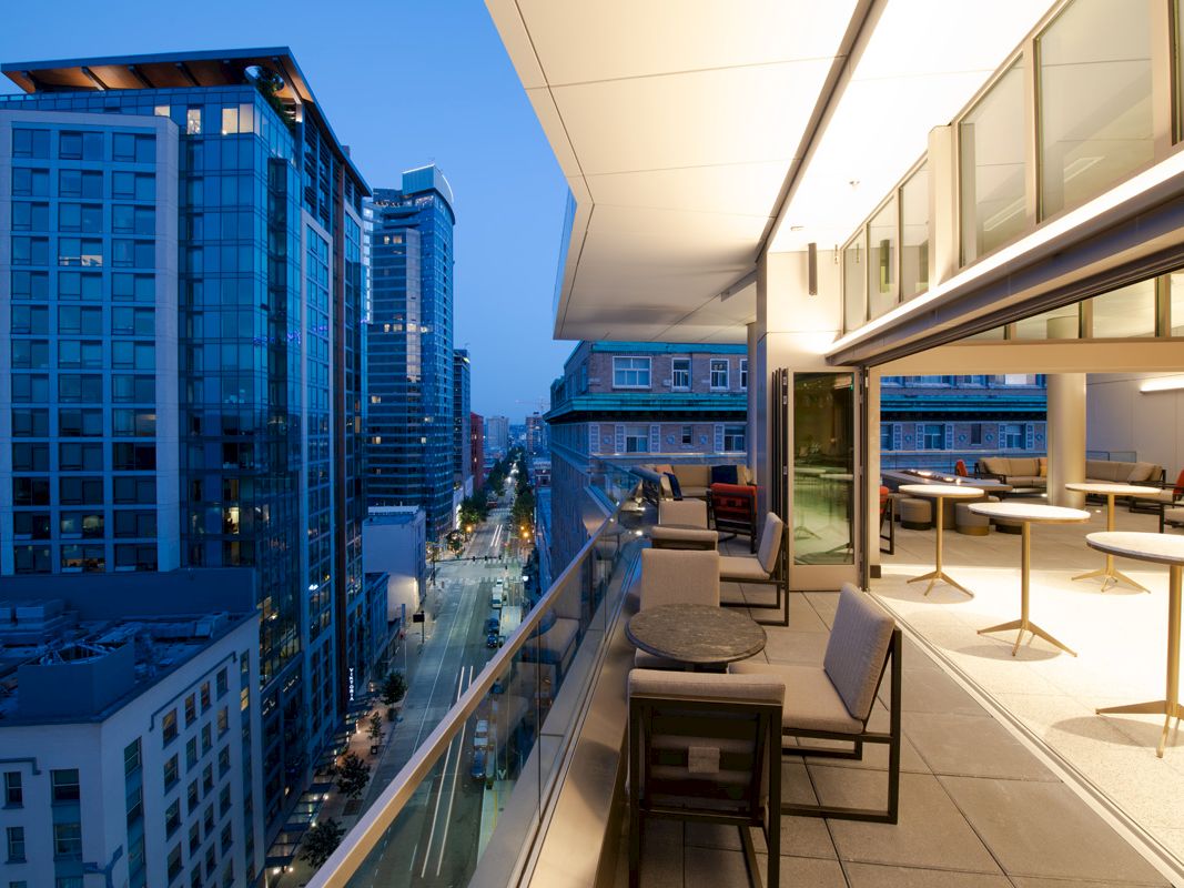 The image shows a modern outdoor balcony with seating, overlooking a city street with high-rise buildings during twilight.
