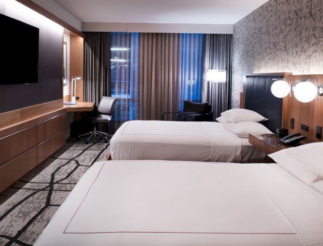The image shows a modern hotel room with two neatly made beds, a flat-screen TV on the wall, a desk, and large windows with curtains.