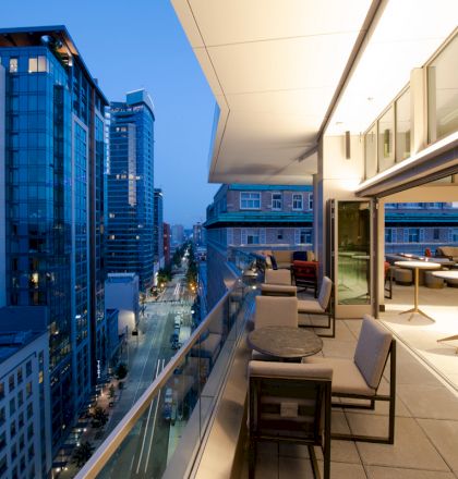 An outdoor terrace with modern seating overlooks a city street lined with tall buildings, illuminated beautifully at dusk.