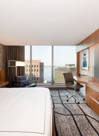 A modern hotel room with a large bed, wall-mounted TV, desk, chair, and a view of city buildings through a large window.