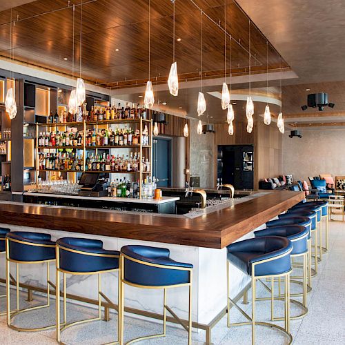 The image shows a modern bar area with a well-stocked bar, blue cushioned stools, hanging lights, and a stylish seating area in the background.