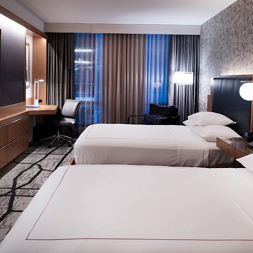 The image shows a modern hotel room with two beds, a flat-screen TV, a desk and chair, and large windows with curtains.