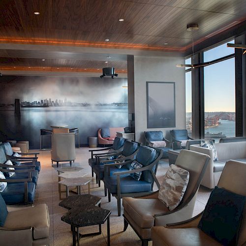A modern lounge area with seating, a city mural, and large windows with views of water and a Ferris wheel, well-lit by natural light.