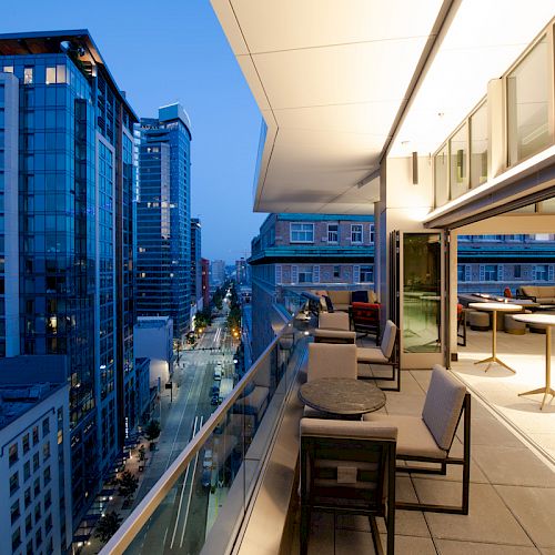 This image shows a modern cityscape at dusk with high-rise buildings and a spacious, stylish outdoor terrace with seating and tables.