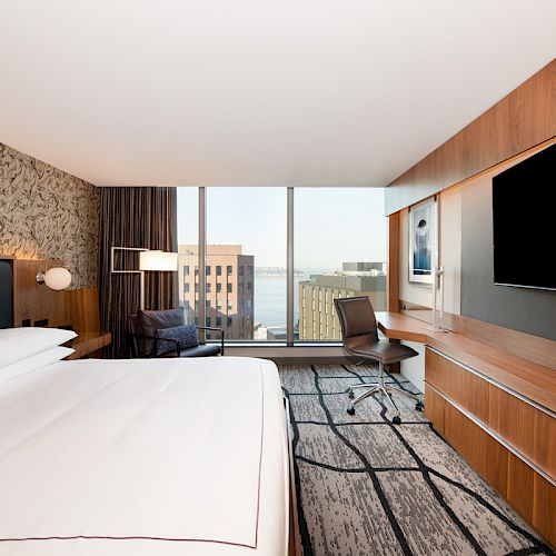 A modern hotel room with a large bed, wall-mounted TV, desk, chair, and a window offering a view of city buildings and the water.