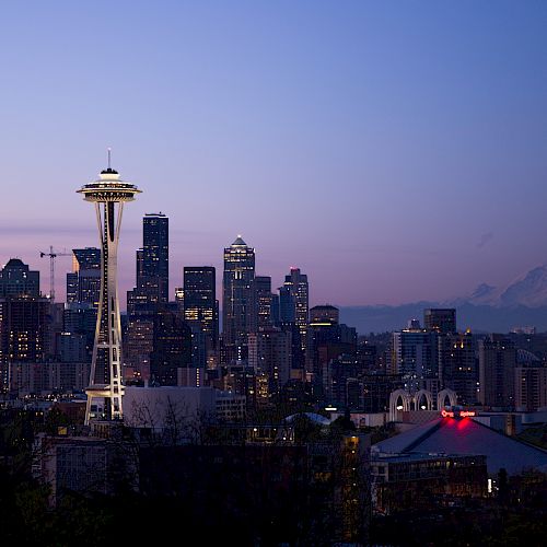 A cityscape at dusk showing a prominent tower, skyscrapers, and a mountain in the background under a fading blue and purple sky.