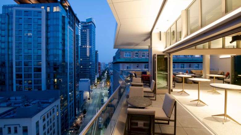 A modern city scene at dusk featuring a rooftop terrace with seating, tables, and a view of the surrounding high-rise buildings and illuminated street.