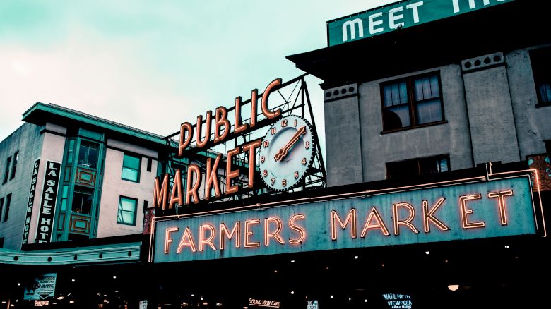 The image shows a public market and farmers market sign with a clock, alongside a building and a partial view of another structure.